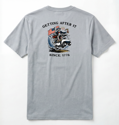 Back of Light gray T-shirt with Uncle Sam Skeleton Character in a Golf Cart carrying an American Flag with Text surrounding that says Getting After it Since 1776