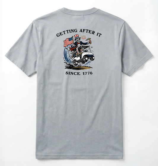 Back of Light gray T-shirt with Uncle Sam Skeleton Character in a Golf Cart carrying an American Flag with Text surrounding that says Getting After it Since 1776