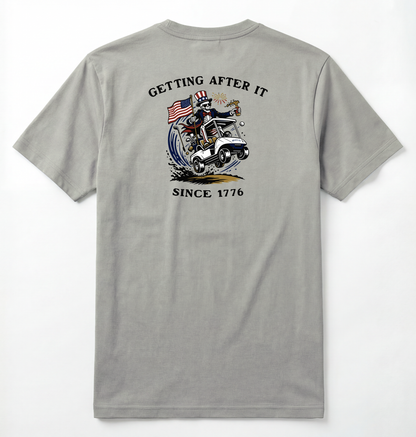 Back of Light gray T-shirt with Uncle Sam Skeleton Character in a Golf Cart carrying an American Flag with Text surrounding that says Getting After it Since 1776