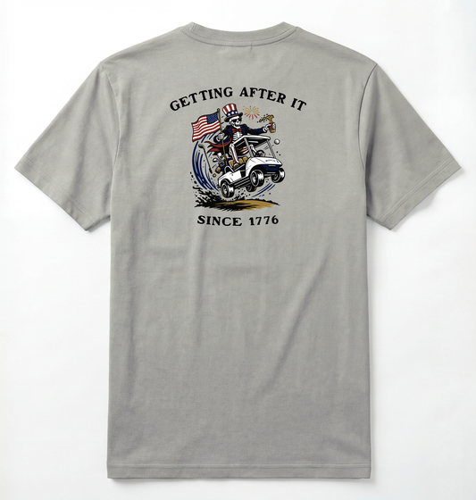 Back of Light gray T-shirt with Uncle Sam Skeleton Character in a Golf Cart carrying an American Flag with Text surrounding that says Getting After it Since 1776